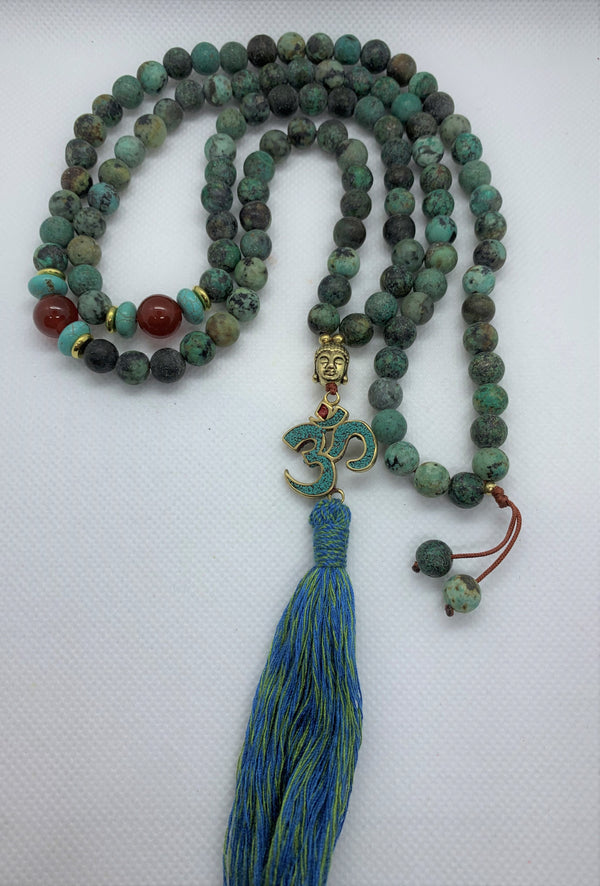 The Evolution Mala with African Turquoise and Carnelian beads necklace features a Buddha & Om symbol pendant, symbolizing the persistence and teachings of Buddha and the most fundamentally strong sound of the universe - Om.