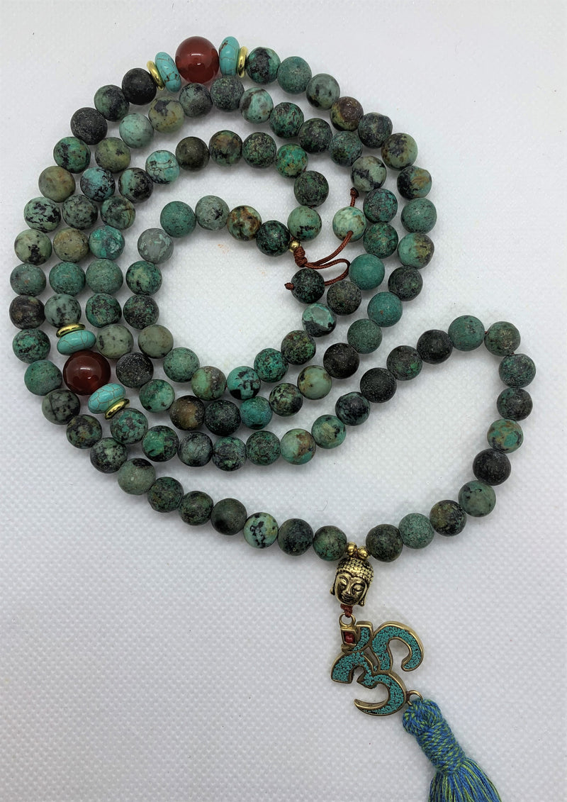 The Evolution Mala with African Turquoise and Carnelian beads necklace features a Buddha & Om symbol pendant, symbolizing the persistence and teachings of Buddha and the most fundamentally strong sound of the universe - Om.
